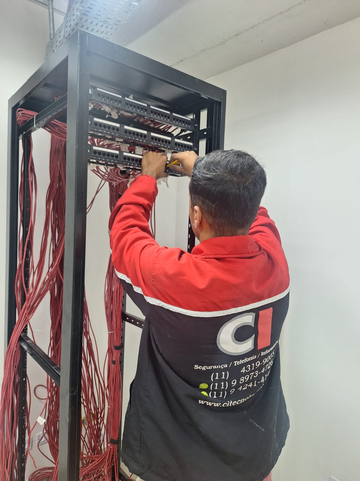 Técnico com jaqueta CI Tecnologia trabalhando em rack de rede com cabeamento estruturado